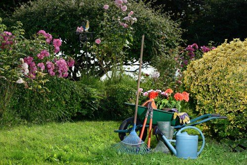 The image depicts a well-maintained backyard garden with a lush, green lawn in the foreground. Surrounding the lawn are various flowering shrubs and bushes, predominantly with pink and purple blossoms, creating a vibrant, colourful backdrop. In the middle of the scene, gardening tools such as a rake, a watering can, and a wheelbarrow filled with supplies are arranged on the grass, indicating active garden maintenance. Behind these tools, a small wooden stake supports a flowering plant or shrub. The background includes taller trees and dense foliage, providing privacy and shade, while the overall scene is lit by natural daylight, suggesting a clear, sunny day. The garden appears to be part of a residential property in Mortlake, with a natural and inviting outdoor space that benefits from ongoing care by gardening professionals from Gardening Services Mortlake, who ensure the healthy growth and presentation of the garden elements. The environment reflects a peaceful, well-kept outdoor area suitable for leisure and outdoor activities within a suburban setting near the Thames suburbs in London postcode area.