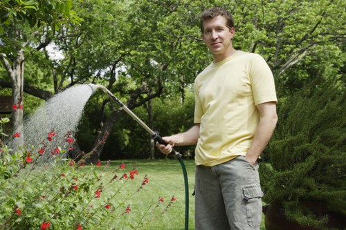 A man wearing a light yellow t-shirt and grey cargo shorts is standing in a lush, well-maintained garden in Mortlake, holding a garden hose and watering red flowering plants in a bed bordered by a curved edge. Behind him, the garden features a neatly trimmed lawn with vibrant green grass, surrounded by various mature trees and bushy shrubs that provide a natural backdrop. To the right, there is a dense green hedge, while in the background, a tall, leafy tree offers partial shade and a natural environment. The scene appears to be taken on a bright, partly cloudy day, with diffuse sunlight illuminating the garden's diverse textures and colours, demonstrating typical outdoor greenery services that enhance garden aesthetics and health.