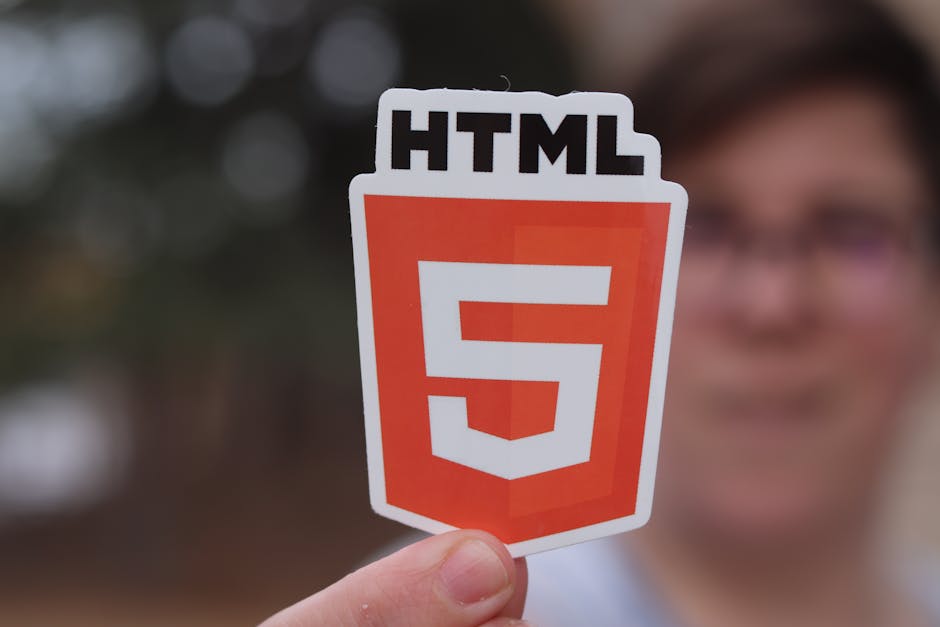 A person holding a sticker featuring the HTML5 logo, which displays a stylized orange and white shield with the number 5 in the center and the text 'HTML' at the top. The background is blurred, includ